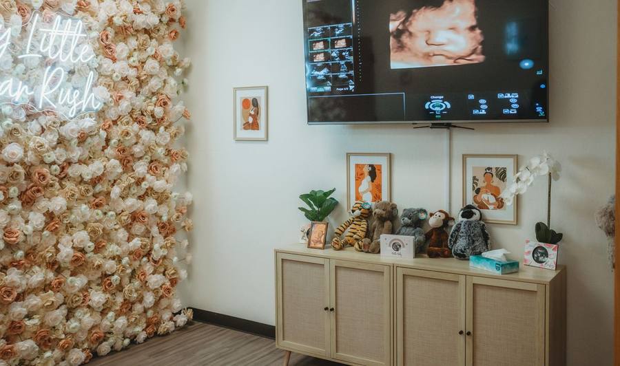 The Sweet 3D Baby scan room with a flower wall, neon sign, wall-mounted screen showing a 3D baby face, and a shelf of heartbeat plushies.